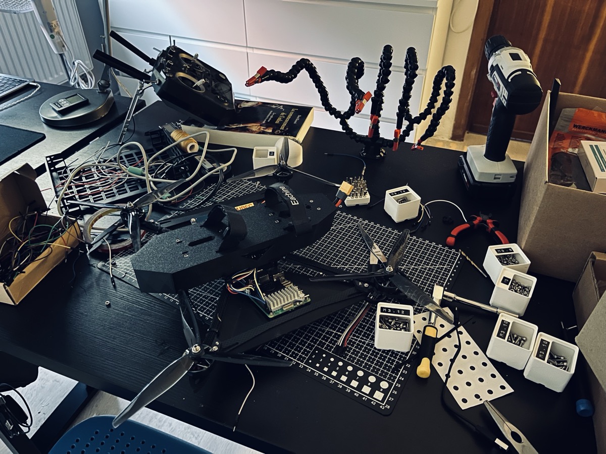 A cluttered workbench mid-build: a drone frame with propellers, a flight controller, screw organizers and tools